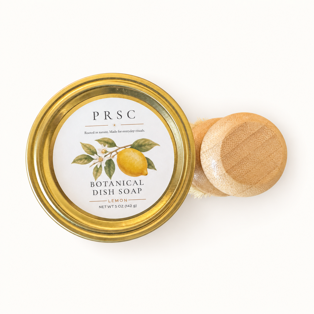 PRSC Botanical Dish Soap in a clear glass jar with a gold metal lid, shown next to a small wooden-handled dish brush on a clean white background.