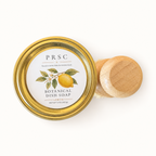 PRSC Botanical Dish Soap in a clear glass jar with a gold metal lid, shown next to a small wooden-handled dish brush on a clean white background.