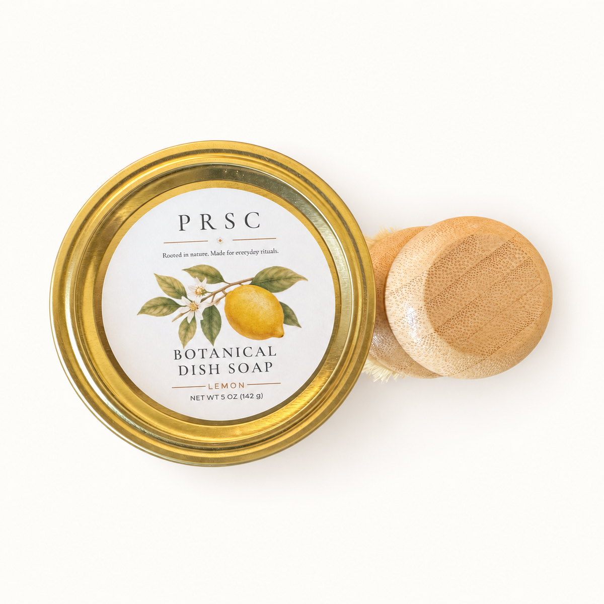 PRSC Botanical Dish Soap in a clear glass jar with a gold metal lid, shown next to a small wooden-handled dish brush on a clean white background.