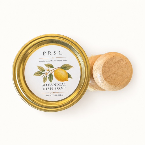 PRSC Botanical Dish Soap in a clear glass jar with a gold metal lid, shown next to a small wooden-handled dish brush on a clean white background.